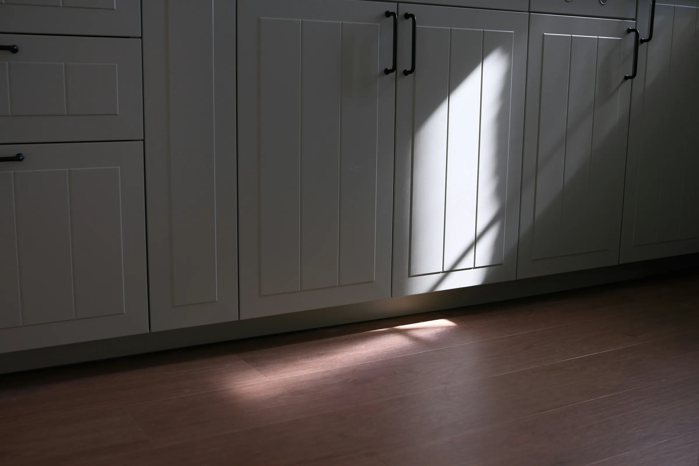 Stock photo: white paneled lower cabinet doors and drawer fronts with black bar pulls, crossed by a slant of sunlight above a brown wood-look floor